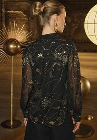 Blusa nera trasparente con motivo celestiale dorato, caratterizzata da stelle e lune. Polsi con bottoni e un colletto classico completano il design.