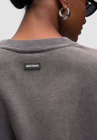 Sudadera gris con una textura suave, que presenta un cuello acanalado y una etiqueta rectangular negra cosida en la parte posterior con el texto "WRSTBHVR."