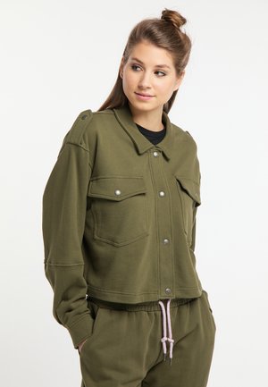 Junge Frau mit braunem Haar im Dutt trägt eine olivgrüne Jacke mit Knöpfen und passende Hose mit pinker Kordel, sie schaut zur Seite.