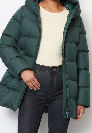 Person iført en mørkegrøn dunjakke over en cremefarvet langærmet top og mørkeblå, højtaljede jeans, der står med det ene hånd i lommen.