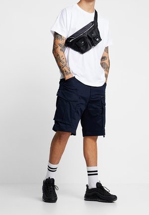 Hombre con camiseta blanca, pantalones cortos de carga negros, zapatillas negras, calcetines blancos con rayas, bandolera negra y tatuajes en los brazos, de pie con las manos en los bolsillos.