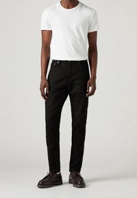 Vaqueros de algodón negros con un corte ajustado, diseño de cinco bolsillos y botones metálicos, combinados con una camiseta blanca lisa de manga corta y zapatos marrones.