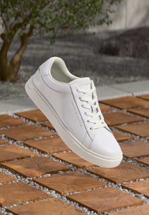 Sneaker en cuir blanc avec des accents texturés, des lacets plats et un design minimaliste. Présente un branding subtil sur le côté.