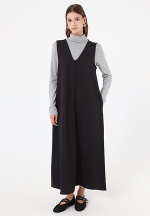 Mujer que lleva un vestido largo negro sin mangas sobre un jersey de cuello alto gris claro con zapatos planos negros sobre un fondo blanco liso.