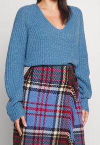 Sininen neulepusero, jossa on ribbikuvio ja V-kaula-aukko, yhdistettynä ruutuiseen hameeseen, jossa on punaista, vihreää ja sinistä sävyä, kiinnitetty mustilla metalliosilla.