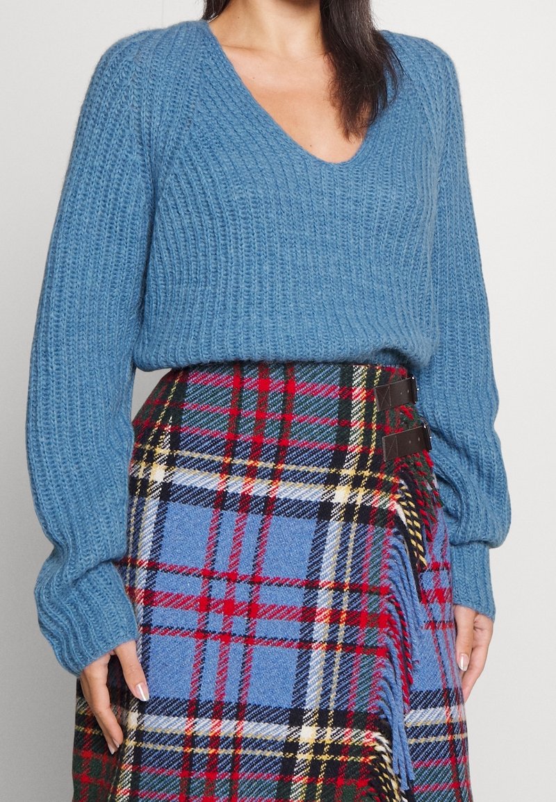 Sininen neulepusero, jossa on ribbikuvio ja V-kaula-aukko, yhdistettynä ruutuiseen hameeseen, jossa on punaista, vihreää ja sinistä sävyä, kiinnitetty mustilla metalliosilla.