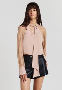 Frau in einer blassrosa Bluse mit Spitzenärmeln und Halsband, kombiniert mit schwarzen Leder-Shorts und goldenen Ringen.