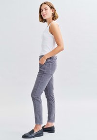 Grijze taps toelopende broek van zachte stof, gecombineerd met een mouwloos wit topje. De broek heeft voorzakken en een zachte textuur. Zwarte loafers.