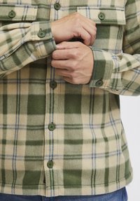 Groene en beige geruite flanellen overhemd met twee borstzakken, knoopsluiting en groene knopen, gemaakt van een zachte, gestructureerde stof.