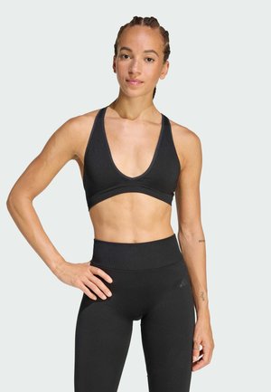 Fit woman with braided hair wears black sports bra and high-waisted leggings, standing with one hand on hip against plain background.