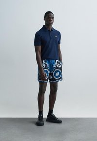 Μπλε ναυτικό κοντομάνικο πόλο συνδυασμένο με μπλε σορτς με μοτίβο που περιλαμβάνει λευκές και μαύρες λεπτομέρειες. Το μοντέλο φοράει μαύρα αθλητικά παπούτσια.