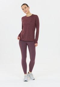 Bordeaux långärmad topp med rund halsringning, kombinerad med tajta plommonleggings. Modellen har på sig grå träningsskor framför en vit bakgrund.