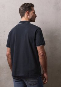 Homme portant un polo bleu marine foncé avec des bordures blanches au col et aux manches, debout contre un mur en béton gris clair, tourné de dos.
