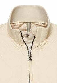 Beżowy, pikowany kołnierz kurtki z częściowo zasuniętym czarnym zamkiem błyskawicznym oraz prążkowanym, wewnętrznym kołnierzem na jednolitym tle.