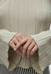 Lys grønn, plisektert topp med utsvingte ermer. Stoffet har en myk tekstur med vertikale striper. Hendene er foldet foran.