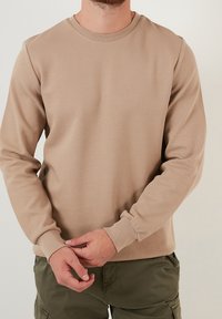 Felpa beige a maniche lunghe con superficie strutturata, scollo tondo e polsini a costine, indossata sopra pantaloni verde oliva.