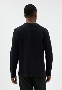 Schwarzes langärmliges Shirt aus strukturiertem Rippstoff, mit einem einfachen Rundhalsausschnitt und lockerer Passform, von hinten gezeigt.