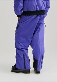 Pantalones de esquí impermeables morados con detalles en negro, con bolsillos con cremallera y un ajuste holgado, combinados con botas de esquí negras.