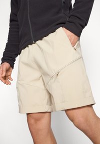Beige shorts vyrobené z lehkého materiálu, s kapsou na zip, ohrnutými manžetami a šikmými švy pro moderní design.