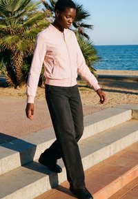 Giovane con giacca rosa e pantaloni neri che scende gradini di pietra vicino a palme e al mare in una giornata soleggiata.
