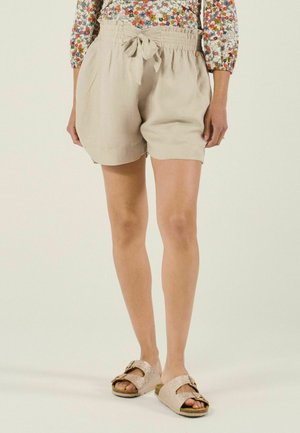 Shorts beige à taille paperbag avec un nœud à l'avant, portés avec des sandales à talons dorées scintillantes et un haut à manches longues à motifs floraux.