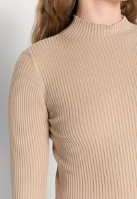 Beżowy sweter z wysokim kołnierzem, wykonany z ribbowanego włókna. Tekstura ma pionowe linie, a materiał wydaje się miękki i dopasowany.