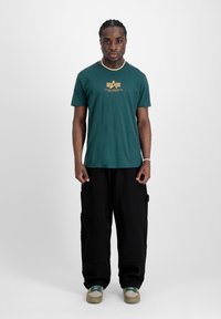 Zelené bavlněné tričko s logem a žlutým designem, v kombinaci s volnými černými cargo kalhotami a teniskami se zelenými akcenty. Jednoduchý, ležérní outfit.