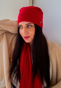 Berretto di lana rosso ben posizionato sulla testa, abbinato a una sciarpa rossa coordinata. Lo sfondo presenta un tessuto beige con texture.