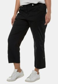 Pantalons noirs coupés avec une texture lisse, deux poches avant et des détails en bouton argenté, associés à des baskets blanches.