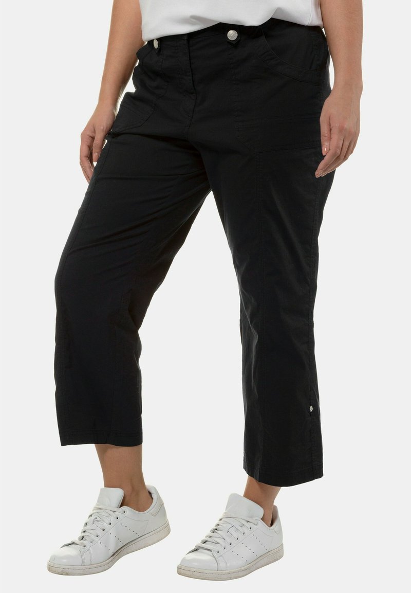 Zwarte cropped broek met een gladde textuur, twee voorkantenzakken en zilveren knoopdetails, gecombineerd met witte sneakers.