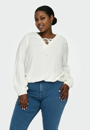 Biała bluzka z dekoltem w kształcie litery V i koronkowym detalem, z bufiastymi rękawami. Połączona z ciemnoniebieskimi dżinsami. Gładka tekstura materiału.