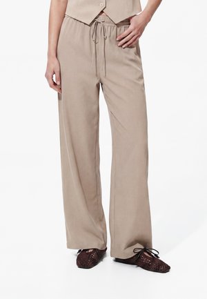 Personne portant un pantalon beige ample à cordon et des chaussures plates marron tressées, la main posée sur la hanche, sur fond blanc.