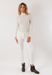 Maglione a maniche lunghe, colore crema, con collo alto e texture liscia, abbinato a pantaloni bianchi slim-fit e stivaletti neri alla caviglia con cerniere.