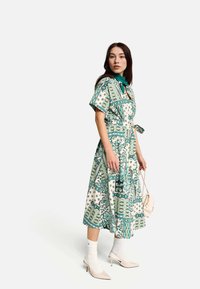 Šaty s květinovým potiskem v zelené a krémové barvě, krátké rukávy, stažené v pase a s páskem na zavázání. Doplňují je bílé ponožky a béžové podpatky.