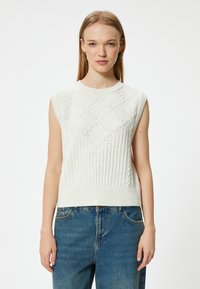 Sweat à mailles sans manches de couleur blanche, arborant un motif en diamant texturé et un ourlet côtelé. Associé à un jean en denim bleu.