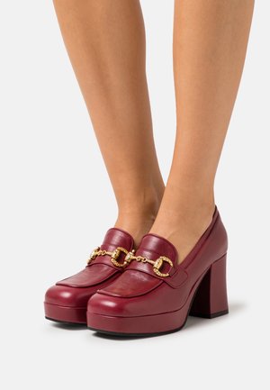 Talons compensés en cuir bordeaux avec détail en chaîne dorée portés pieds nus sur fond blanc.