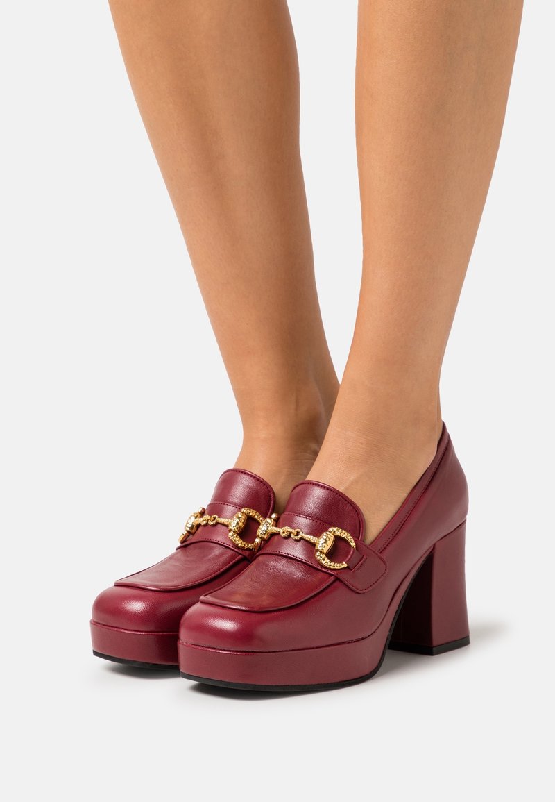 Mørkerøde læder platformhæle med guld kædelynkedetalje båret på baræ fødder mod en hvid baggrund.