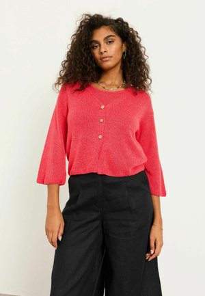 Woman with curly hair wearing a coral pink buttoned sweater and black wide-leg pants, standing against a plain light background.