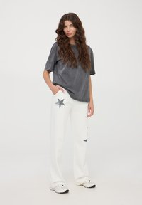 Szara bawełniana koszulka o luźnym kroju i krótkich rękawach, zestawiona z białymi spodniami z ciemnymi wzorami w gwiazdy, noszona z białymi sneakersami.