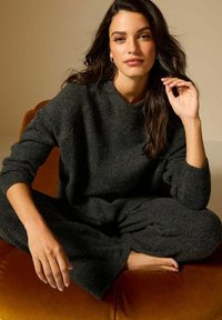 Completo in maglia grigio scuro, composto da un maglione a girocollo e pantaloni, con vestibilità rilassata, texture morbida e maniche lunghe, indossato mentre si è seduti su una superficie arancione.
