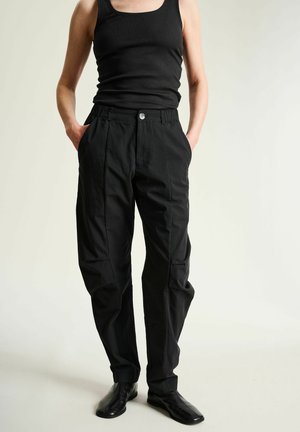 Persona con camiseta sin mangas negra, pantalones negros de cintura alta con bolsillos y zapatos negros, de pie contra un fondo claro y liso.