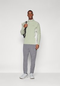 Haut en polaire vert clair à fermeture éclair demi-longue avec des panneaux latéraux blancs, associé à un pantalon de randonnée gris. Le modèle se tient en tenant un sac à dos. Chaussures de sport blanches.