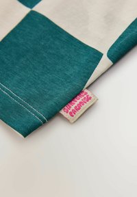 Bombažno blago z vzorcem karo v barvah modrozeleni in krem. Poudarjeni šivi v kontrastni barvi ter roza etiketa v kotu z belim napisom.