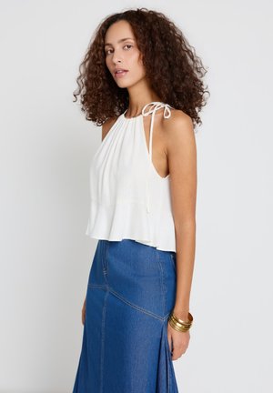 Woman with curly hair wearing a white sleeveless top with tie straps and a blue denim skirt, accessorized with gold bracelets.