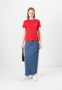 Rød t-skjorte med korte ermer og rund grafikk, kombinert med et langt blått denimskjørt. Modellen har på seg hvite sneakers og holder en svart veske.