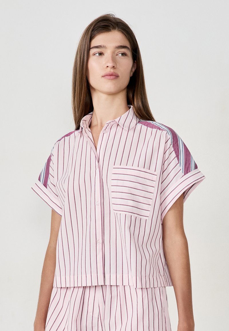 Femme portant une chemise courte à manches avec des rayures roses, dotée d'une poche sur la poitrine, et un pantalon assorti à rayures, regardant vers la droite.