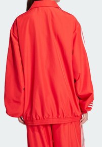 Punainen polyesterinen urheilutakki pystypäisellä kauluksella, löysällä istuvuudella, joustavalla vyötäröllä ja valkoisilla raidoilla hihojen resoreissa.