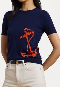 Pull à manches courtes navy avec un motif d'ancre orange. Tissu tricoté, col rond et coupe ajustée. Associé à un pantalon de couleur crème.