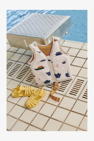 Lightweight children's life jacket with boat patterns, beige color, and front buckle. Yellow rubber sandals nearby. Tiled poolside background.