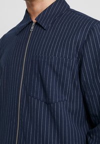 Chaqueta azul marino con rayas finas, elaborada en tela texturizada, con cremallera frontal, cuello y bolsillo en el pecho.